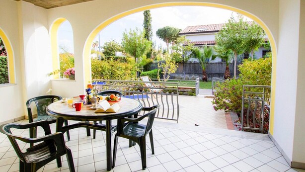 An outdoor patio with a round table and chairs, surrounded by arches and a view of a garden and buildings. - Apartment in Cilento Near Sandy Beach (Ascea)