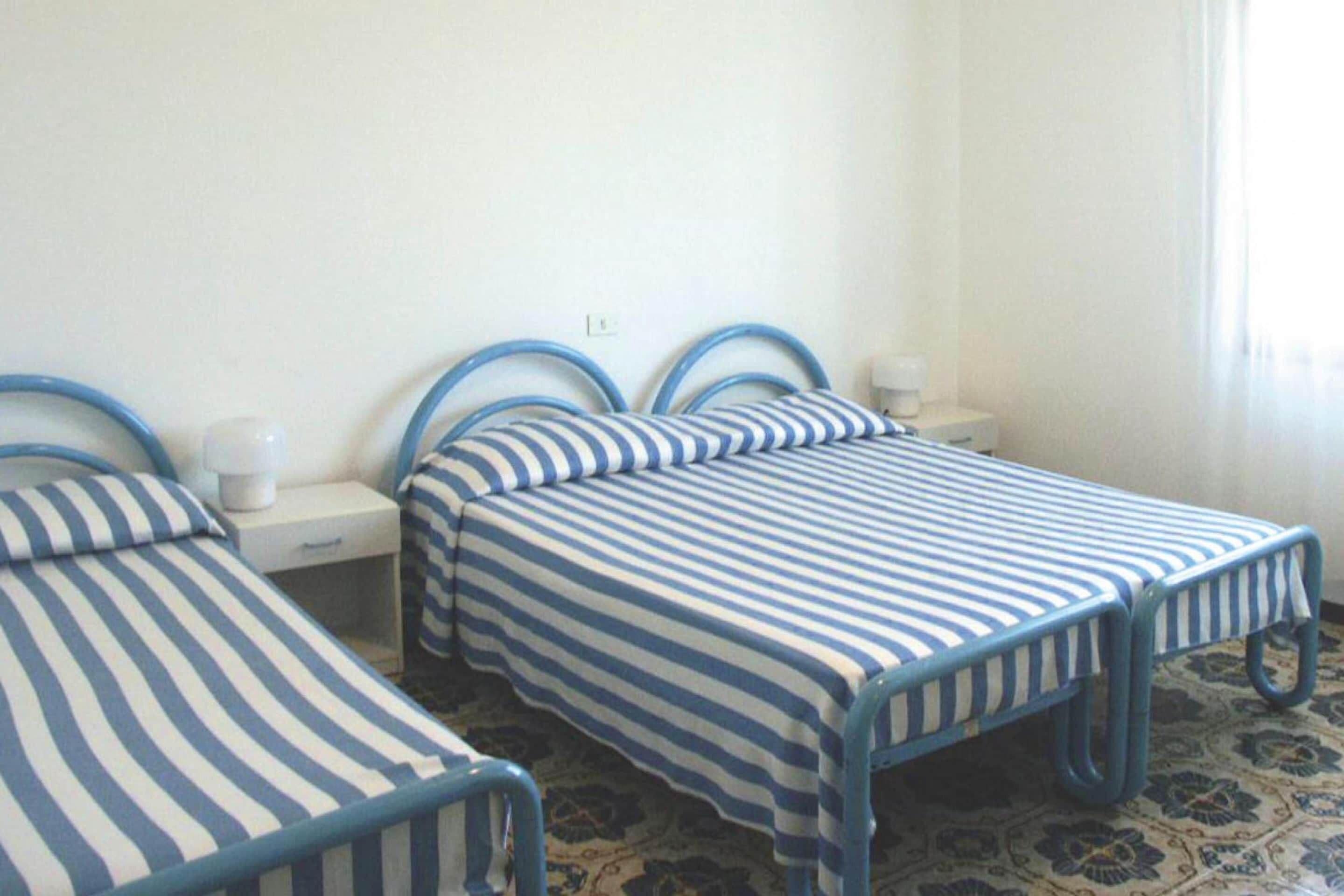 Quarto com duas camas, cada uma com estrutura de metal azul e roupa de cama listrada. Apresenta uma mesa de cabeceira com abajur e uma pequena mesa também com abajur. O chão possui um design de azulejos padronizado.