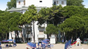 Et hotel ved stranden med flere etager, udendørs siddepladser med parasoller og en legeplads til børn.