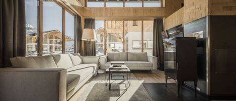 A spacious living room with a high ceiling, wooden beams, and large windows offering a view of the mountains. There is a sofa, a coffee table, and a flat-screen TV.