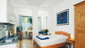 A bedroom with a wooden bed, a nightstand, a wooden wardrobe, a window with a view of flowers, and a kitchenette with a sink and stove. - Studio, Wyk auf Fohr (Wyk auf Foehr)