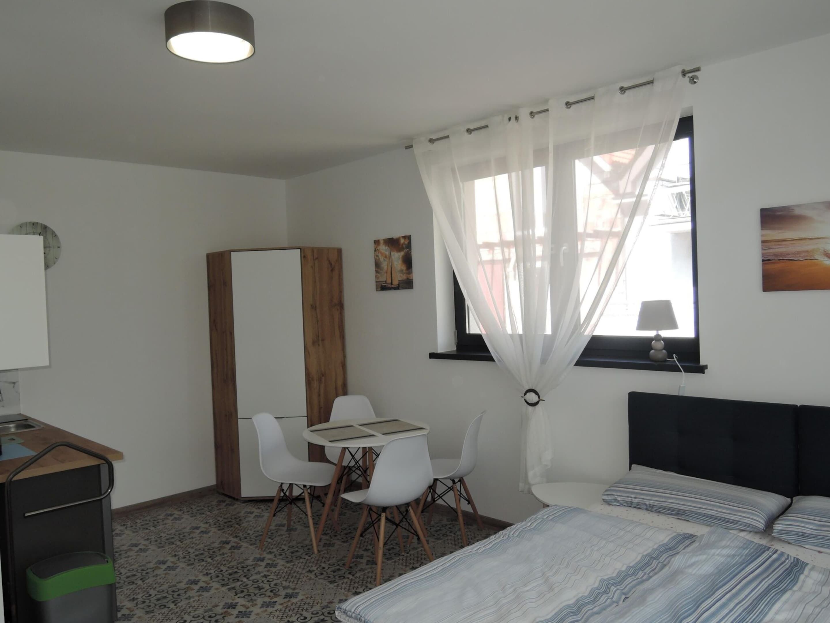 A bedroom with a bed, a desk with chairs, a wardrobe, and a window with sheer curtains.