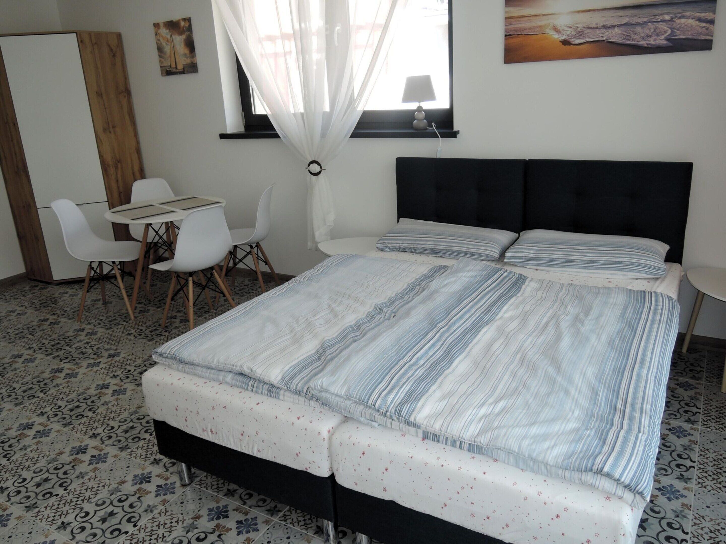 A bedroom with a bed, a desk, two chairs, a window with curtains, and a painting on the wall.
