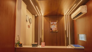 Un espace d’accueil d’hôtel avec un comptoir de réception en bois, des horloges murales, un appareil de climatisation et un petit objet décoratif posé sur le comptoir.