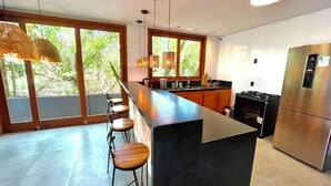 A modern kitchen with a large island, bar stools, a refrigerator, and a view of greenery outside.