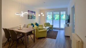 A modern living room with a dining table, green sofas, and a large window with a view of greenery. - Ferienwohnung im Dunenpark Binz (Binz)