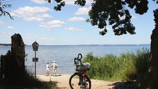 Una bicicleta estacionada en un camino de arena junto a un lago, con un cartel y dos personas al fondo.