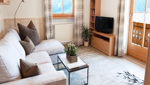 Un soggiorno con un divano, un tavolino da caffè con una pianta, una TV su un mobile in legno e una finestra con tende.