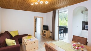 A modern living room with a sofa, a wooden coffee table, a dining table with chairs, a TV, and a balcony with a view of greenery. - Adults Only Apartment With Pool (Sankt Kanzian am Klopeiner See)