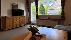 Ein Wohnzimmer mit einem Holzkabinett, einem Fernseher, einem Tisch mit Vase und Fernbedienung sowie einem Fenster mit Vorhängen.