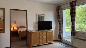 Ein Hotelzimmer mit Holzschrank, Flachbildfernseher, Bett, Nachttisch mit Lampe und einem fenster mit Vorhängen.