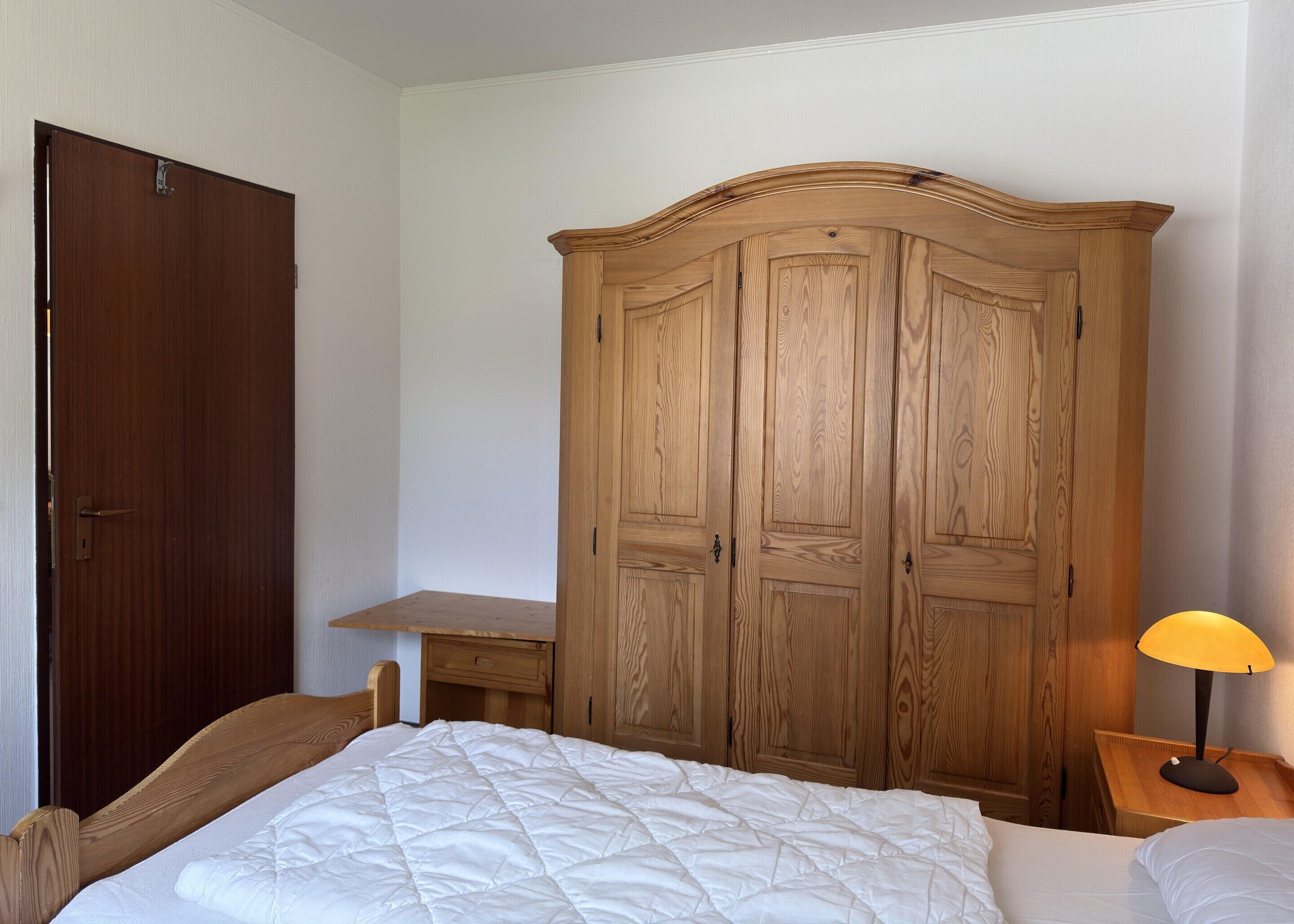 Une chambre à coucher avec une armoire en bois, un lit recouvert d’une couette blanche, une table de chevet avec une lampe et un bureau en bois.