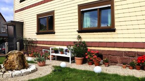A building with a yellow exterior, two windows with brown frames, and a gravel driveway with potted plants and a white bench. - Ferienwohnung, Tambach-dietharz (Tambach-Dietharz)