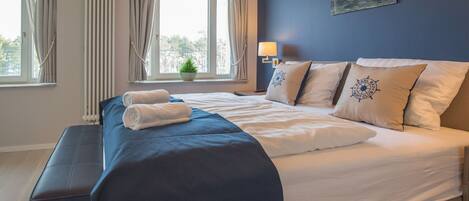 A neatly made bed with white and blue linens, a bedside lamp, and a window with curtains.