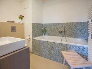 A bathroom with a white bathtub, a wooden bench, a towel rack, and a wall with mosaic tiles.