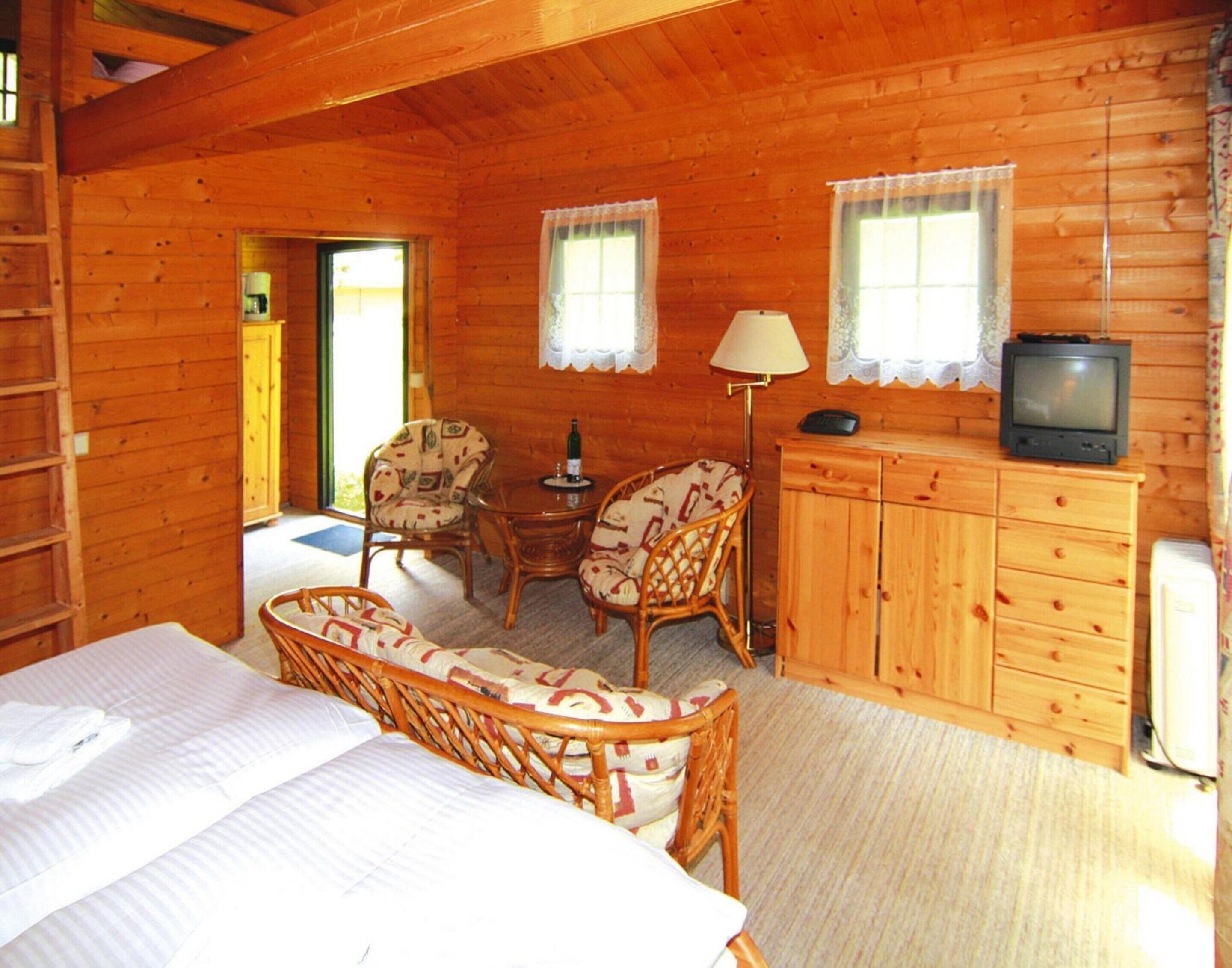 A wooden cabin room with a bed, two wicker chairs, a small table with a bottle, a television on a cabinet, and a refrigerator.