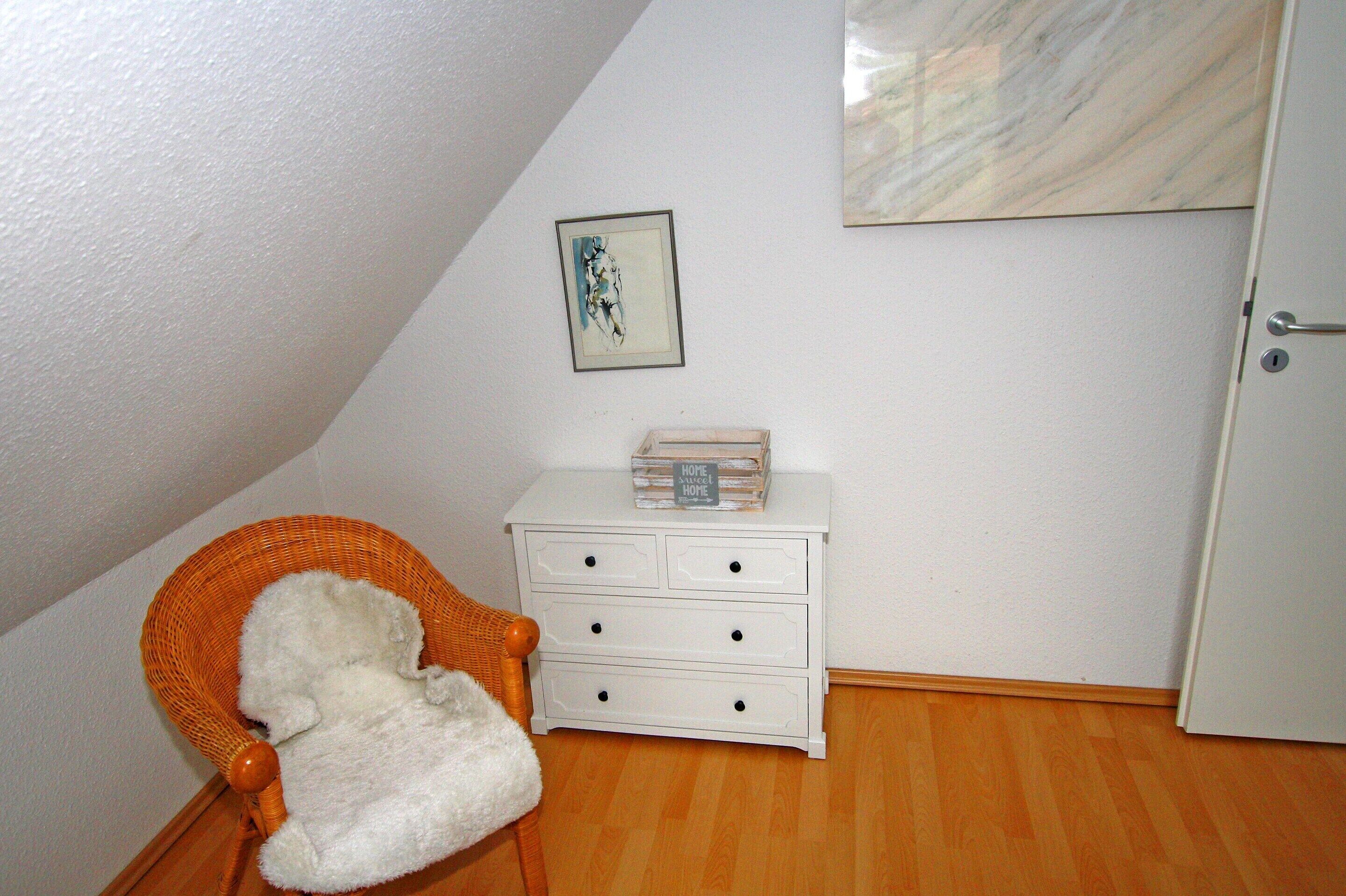 A corner room with a white dresser, a wicker chair with a white cushion, a framed picture on the wall, and a marble-like artwork above the dresser.