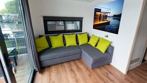 Un salon moderne avec un canapé d’angle gris, des coussins vert vif, un grand téléviseur à écran plat et un tableau encadré représentant une maison au bord du lac.