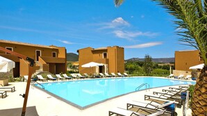 Ein Swimmingpool mit Liegestühlen und Sonnenschirmen, umgeben von Gebäuden vor einer Bergkulisse.