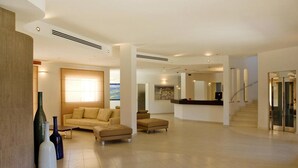 Ein geräumiger Wohnbereich mit einem beigen Sofa, einem Hocker und einem Couchtisch. Auf dem Boden stehen zwei Vasen, an der Wand hängt ein Gemälde und eine Treppe führt zu einer weiteren Ebene.
