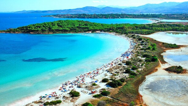 Luftaufnahme einer Küstenlandschaft mit einem langen Strand, gesäumt von Sonnenschirmen und Menschen.