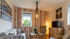 A cozy living room with a striped wall, a sofa, a chair, a table, a TV, and a window with a view of greenery. - Doppelhaushalfte in St. Peter-ording (St. Peter-Ording)