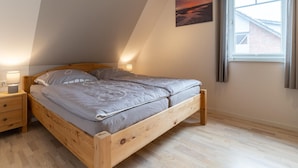 A bedroom with a sloped ceiling, a bed with a grey mattress, wooden headboard, bedside tables with lamps, and a window with curtains. - Doppelhaushalfte in St. Peter-ording (St. Peter-Ording)