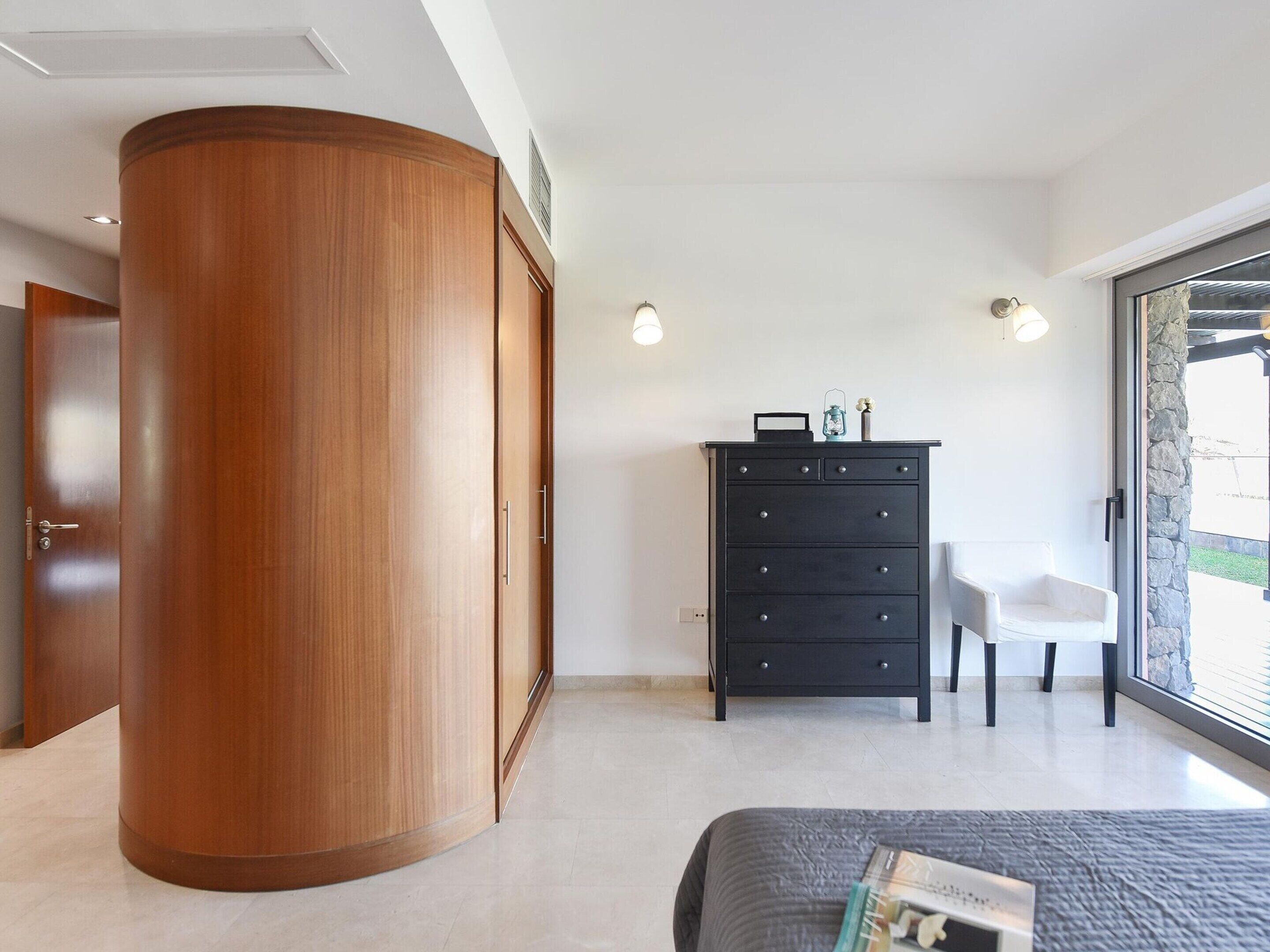 Quarto com guarda-roupa de madeira, cômoda preta, cadeira branca e uma cama.