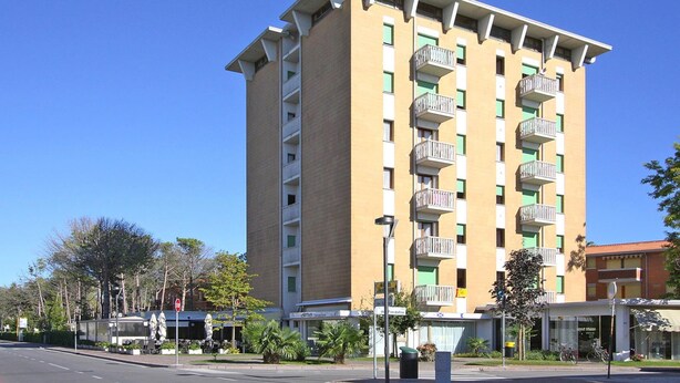 A multi-story hotel with balconies, a clear sky, and a street with parked cars and greenery.