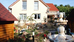 Ein zweistöckiges Haus mit rotem Ziegeldach, einem Garten mit Steinmauer und einer Zierbrunnen, sowie einer Holzkonstruktion auf der linken Seite.