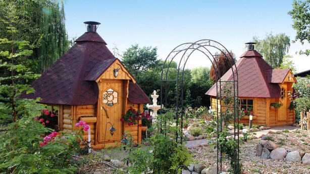 Zwei Holzhütten mit Kegeldächern, umgeben von einem Garten mit Blumen und einem kleinen Springbrunnen.