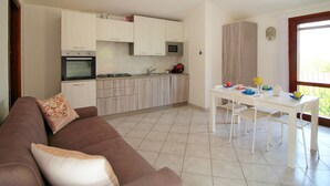 A modern kitchen with white cabinets, a dining table, and a sofa with cushions. - Terraced House in Costa Rei With Garden (Muravera)