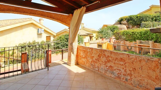 A covered balcony with tiled flooring, a metal railing, and a view of residential buildings and greenery. - Terraced House in Costa Rei With Garden (Muravera)