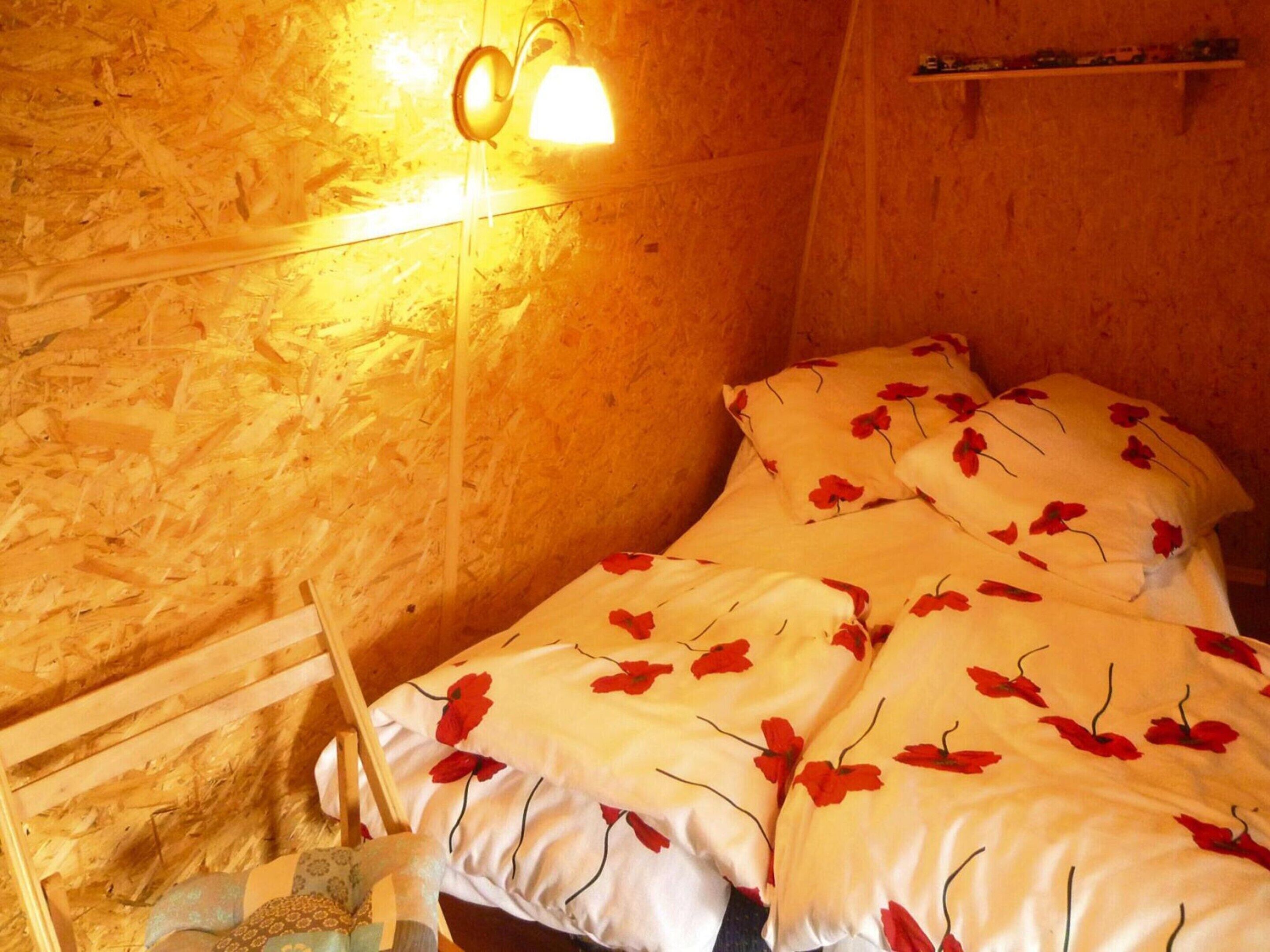 Una habitación con una cama cubierta con ropa de cama blanca y motivos florales rojos, un cabecero de madera y una lámpara de pared.