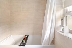 A bathroom with a bathtub, a shower curtain, and a window with a plant on the windowsill. - Charming Haven House Right Next To Moss Bank Park (Bolton)