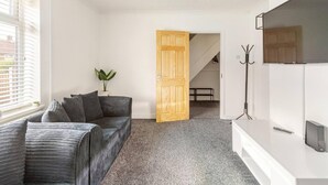 Een moderne woonkamer met een grijze bank, een houten deur, een kapstok en een flatscreen televisie.