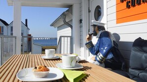 Una terraza de madera con una mesa dispuesta para dos, un muffin y una taza. Se ve una estatua de un marinero a la derecha.