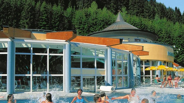Ett poolområde där folk badar och solar, omgivet av en byggnad med stora fönster och en överbyggd sittplats.