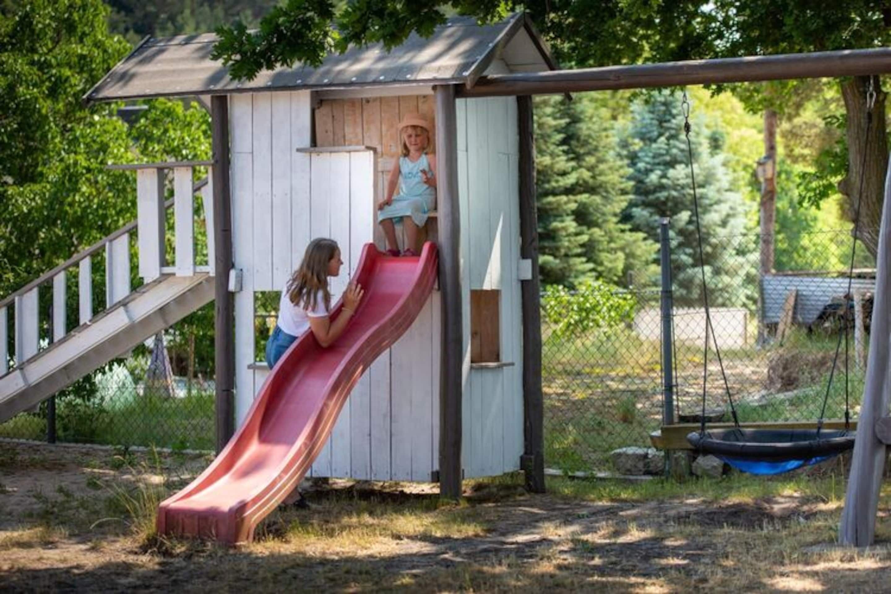 A wooden playhouse with a red slide, a swing set, and a fenced yard.