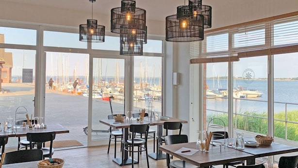 Ein Essbereich mit Tischen und Stühlen, große Fenster mit Blick auf eine Marina und hängende Pendelleuchten.