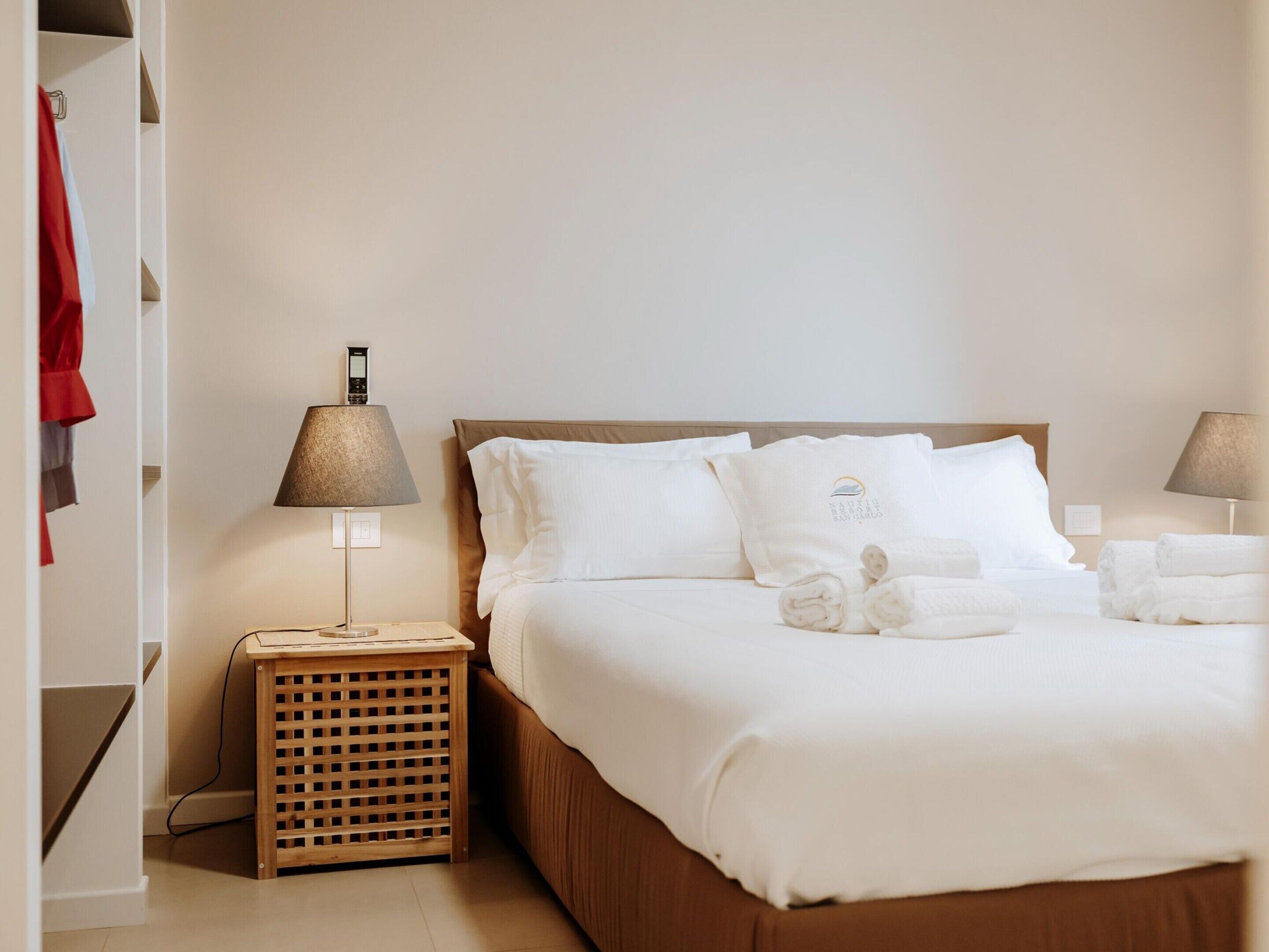 A hotel room with a bed, two bedside tables, a lamp, a small wooden basket, and a red garment hanging on a rack.
