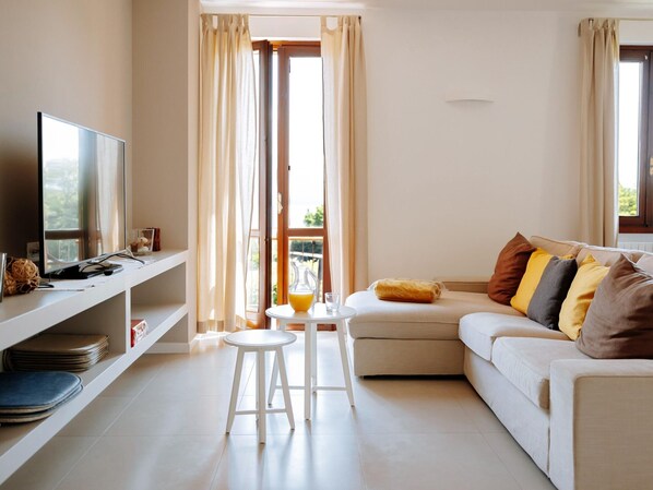 A modern living room with a flat-screen TV, a white sofa with colorful cushions, a small white table with a glass of orange juice, and a white stool. - Il Limone Lake View at Nautic Resort San Carlo (Gargnano)