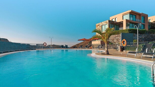 Ein Pool mit Liegestühlen und Sonnenschirmen, ein modernes Gebäude mit großen Fenstern und ein Rettungsring am Poolrand.