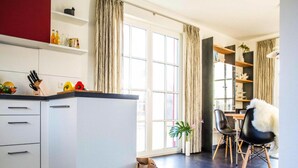 Een moderne keuken met witte kasten, een zwart aanrecht en een raam met gordijnen. Er is een eethoek met een tafel en stoelen, en een tas met boodschappen op de vloer.