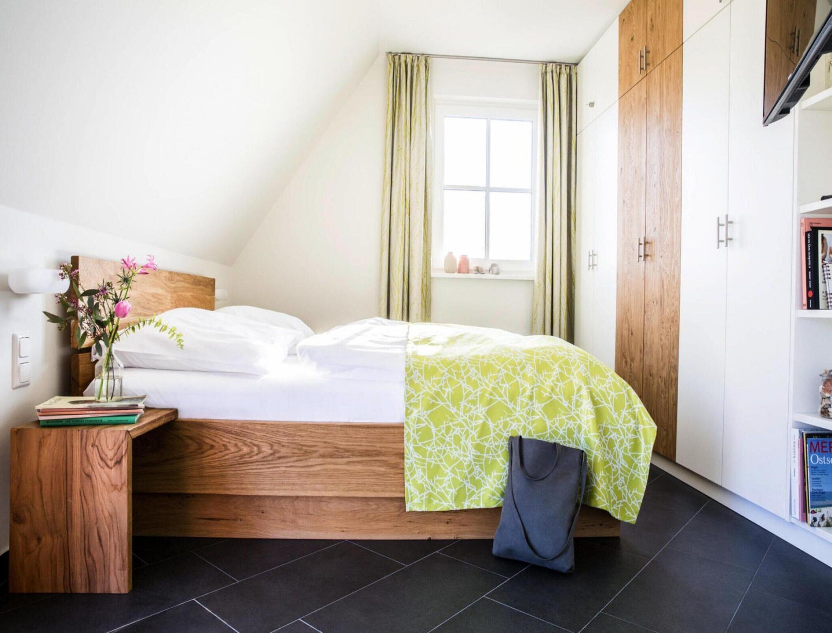 A bedroom with a wooden headboard, a bed with a green patterned bedspread, a bedside table with a vase of flowers, and a bookshelf with books.