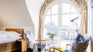 Een slaapkamer met een bed, een nachtkastje, een stoel, een klein tafeltje met een bloevas en een groot raam met gordijnen.