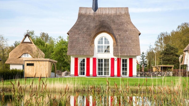Een huis met rieten dak en een groot rondboogvenster, omgeven door een vijver en groen.