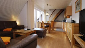 A modern living room with a sofa, coffee table, and TV. There is a kitchen area with wooden cabinets and a dining table. - Appartement in Westerland auf Sylt (Sylt)