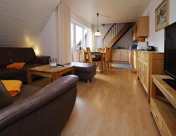 A modern living room with a sofa, coffee table, and TV. There is a kitchen area with wooden cabinets and a dining table. - Appartement in Westerland auf Sylt (Sylt)