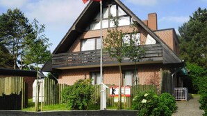 A two-story building with a pitched roof, brick walls, and a flagpole with a flag. There are signs on the fence and a gate leading to the entrance. - Appartement in Westerland auf Sylt (Sylt)